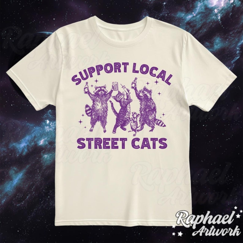 May include: White t-shirt with a purple graphic of three raccoons and a cat holding drinks. The text "Support Local Street Cats" is printed in purple below the graphic.