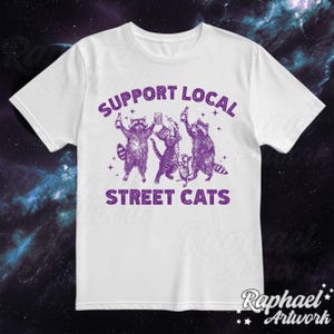 May include: White t-shirt with a purple graphic of raccoons holding drinks and the text "Support Local Street Cats".