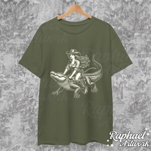 K&ouml;nnte beinhalten: Ein gr&uuml;nes T-Shirt mit einem wei&szlig;en Aufdruck einer Frau im Cowboyhut, die auf einem Alligator reitet.