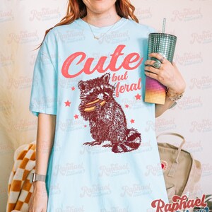 May include: Light blue t-shirt with the text "Cute but Meral" in red script above a raccoon graphic. The raccoon is brown and white, holding a snack. Red stars are scattered around the graphic. The shirt is a casual, unisex style.