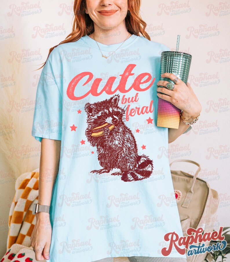May include: Light blue t-shirt with the text "Cute but Meral" in red script above a raccoon graphic. The raccoon is brown and white, holding a snack. Red stars are scattered around the graphic. The shirt is a casual, unisex style.
