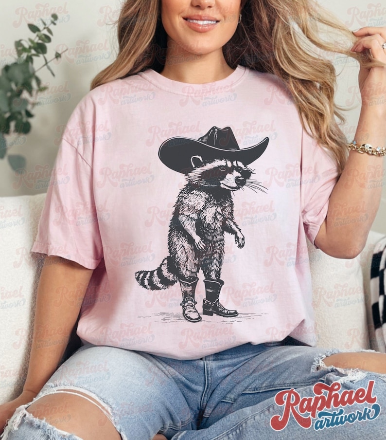 May include: A pink t-shirt with a black and white illustration of a raccoon wearing a cowboy hat and boots. The raccoon is standing on its hind legs and has a serious expression on its face.