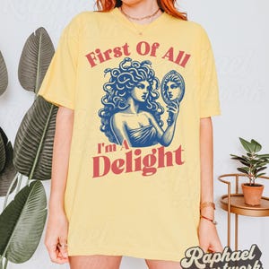 May include: A pale yellow t-shirt featuring a blue graphic illustration of Medusa holding a hand mirror. The text "First Of All" appears in red above the graphic, and the text "I'm A Delight" appears in red below the graphic.