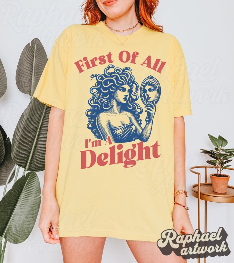 May include: A pale yellow t-shirt featuring a blue graphic illustration of Medusa holding a hand mirror. The text "First Of All" appears in red above the graphic, and the text "I'm A Delight" appears in red below the graphic.