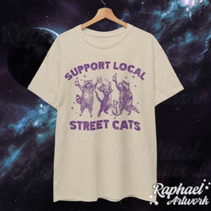 May include: A beige t-shirt with a purple graphic of three raccoons holding drinks and the text "Support Local Street Cats".