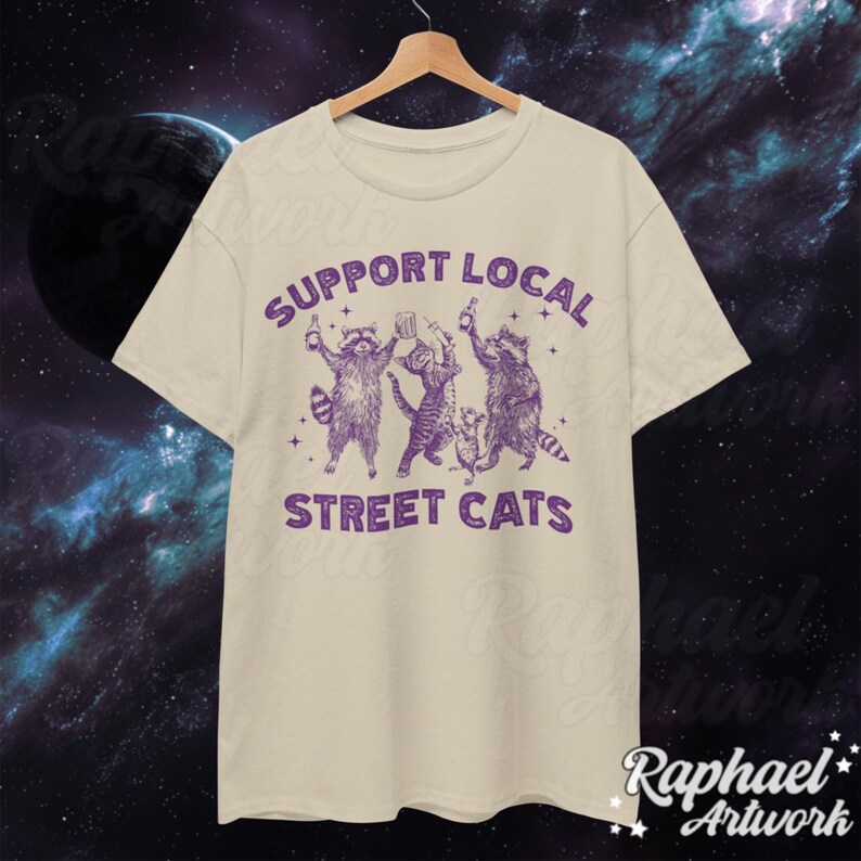 May include: A beige t-shirt with a purple graphic of three raccoons holding drinks and the text "Support Local Street Cats".