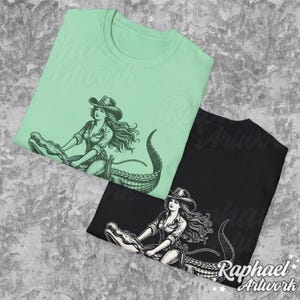 K&ouml;nnte beinhalten: Zwei T-Shirts mit einem grafischen Aufdruck einer Frau, die auf einem Alligator reitet. Die Frau tr&auml;gt einen Cowboyhut und eine Weste. Der Alligator ist schwarz-wei&szlig; auf einem schwarzen T-Shirt und gr&uuml;n auf einem hellgr&uuml;nen T-Shirt.