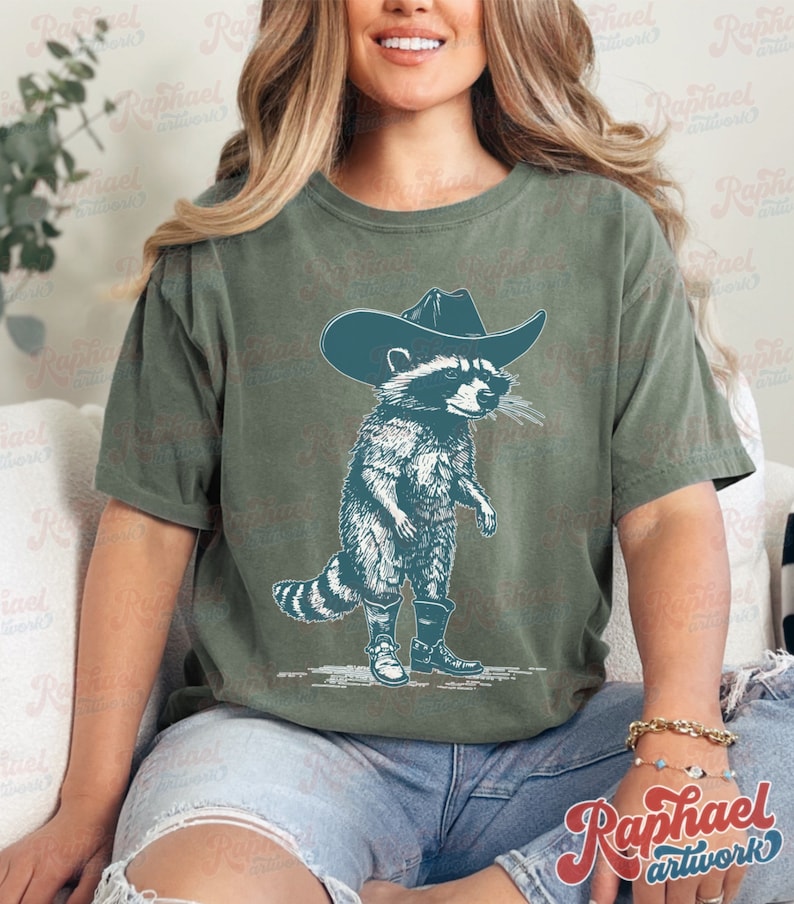 May include: A green t-shirt with a graphic of a raccoon wearing a cowboy hat and boots. The raccoon is standing on its hind legs and has a mischievous expression on its face.