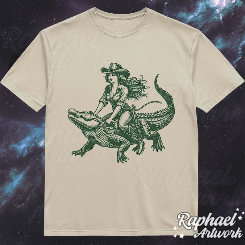 K&ouml;nnte beinhalten: Ein beiges T-Shirt mit einem gr&uuml;nen Grafikdruck einer Frau, die einen Cowboyhut tr&auml;gt und auf einem Alligator reitet.
