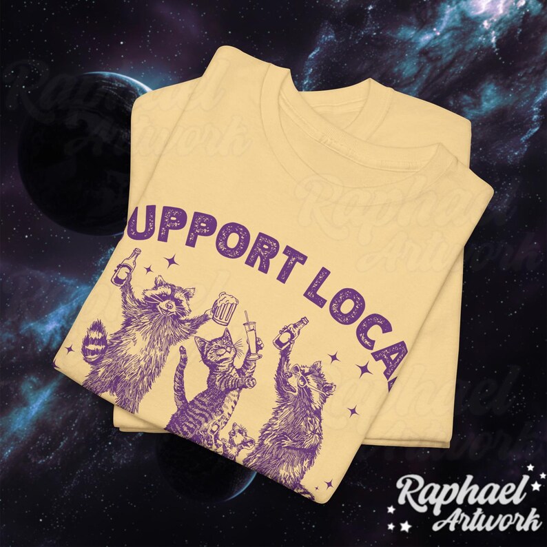 May include: A yellow t-shirt with the text "Support Local" in purple lettering. The shirt features a graphic of three animals, a raccoon, a cat, and another raccoon, all holding drinks. The animals are also in purple.