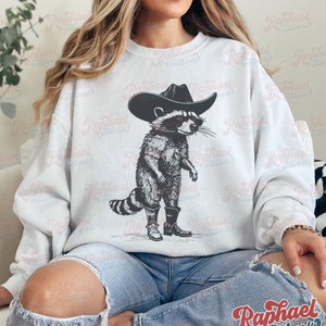 May include: A white sweatshirt with a black and white illustration of a raccoon wearing a cowboy hat and boots. The raccoon is standing with its arms crossed and has a serious expression on its face.