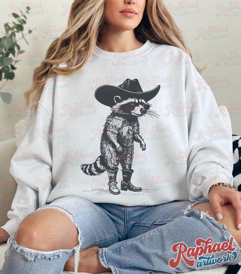 May include: A white sweatshirt with a black and white illustration of a raccoon wearing a cowboy hat and boots. The raccoon is standing with its arms crossed and has a serious expression on its face.