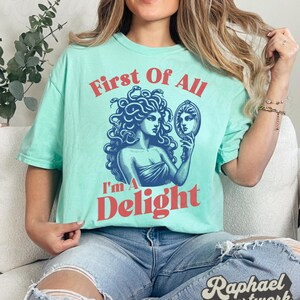 May include: A light mint green short-sleeve t-shirt featuring a dark blue graphic of Medusa holding a mirror. The text "First Of All" appears in red above the graphic, and the text "I'm A Delight" appears in red below the graphic.