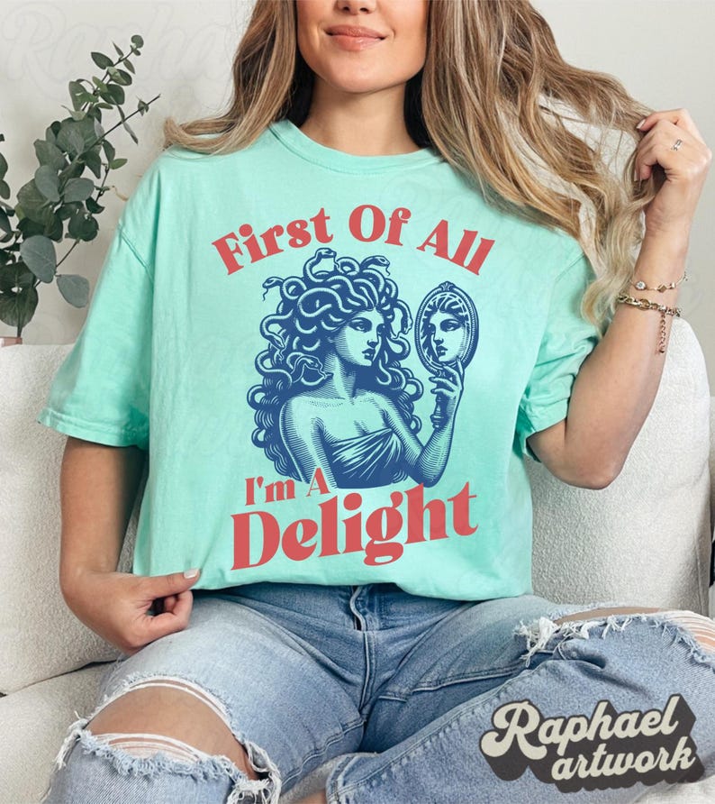 May include: A light mint green short-sleeve t-shirt featuring a dark blue graphic of Medusa holding a mirror. The text "First Of All" appears in red above the graphic, and the text "I'm A Delight" appears in red below the graphic.