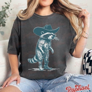 May include: A dark gray t-shirt with a graphic of a raccoon wearing a cowboy hat and boots. The raccoon is standing on its hind legs and has a mischievous expression on its face.