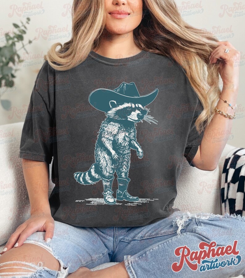 May include: A dark gray t-shirt with a graphic of a raccoon wearing a cowboy hat and boots. The raccoon is standing on its hind legs and has a mischievous expression on its face.