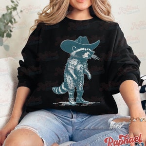May include: A black sweatshirt with a graphic of a raccoon wearing a cowboy hat and boots. The raccoon is standing on its hind legs and has a mischievous expression on its face.