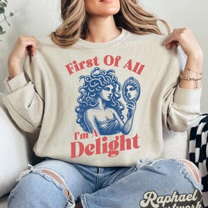 May include: A beige crewneck sweatshirt featuring a blue graphic illustration of Medusa holding a hand mirror. The text "First Of All" is printed in red above the graphic, and the text "I'm A Delight" is printed in red below the graphic.