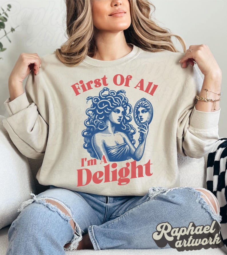 May include: A beige crewneck sweatshirt featuring a blue graphic illustration of Medusa holding a hand mirror. The text "First Of All" is printed in red above the graphic, and the text "I'm A Delight" is printed in red below the graphic.