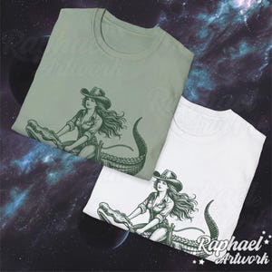 K&ouml;nnte beinhalten: Zwei T-Shirts mit einem grafischen Aufdruck einer Frau, die auf einem Alligator reitet. Die Frau tr&auml;gt einen Cowboyhut und Stiefel. Die T-Shirts sind in Gr&uuml;n und Wei&szlig; erh&auml;ltlich.