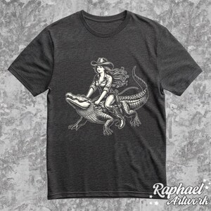 K&ouml;nnte beinhalten: Ein dunkelgraues T-Shirt mit einer Schwarzwei&szlig;illustration einer Frau, die auf einem Alligator reitet. Die Frau tr&auml;gt einen Cowboyhut und Stiefel. Der Alligator blickt nach rechts.
