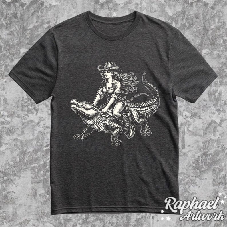K&ouml;nnte beinhalten: Ein dunkelgraues T-Shirt mit einer Schwarzwei&szlig;illustration einer Frau, die auf einem Alligator reitet. Die Frau tr&auml;gt einen Cowboyhut und Stiefel. Der Alligator blickt nach rechts.