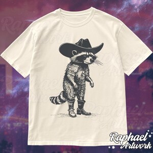 May include: A white t-shirt with a black and white illustration of a raccoon wearing a cowboy hat and boots.