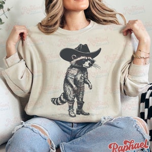 May include: A beige sweatshirt with a graphic print of a raccoon wearing a cowboy hat and boots. The raccoon is standing on its hind legs and has a serious expression on its face.