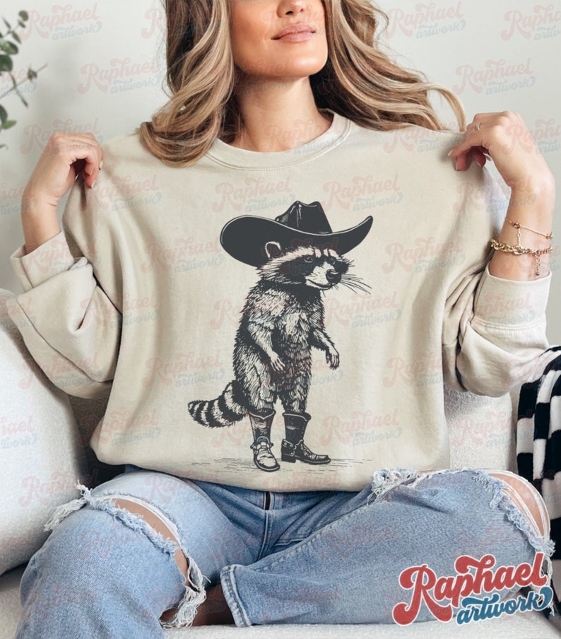May include: A beige sweatshirt with a graphic print of a raccoon wearing a cowboy hat and boots. The raccoon is standing on its hind legs and has a serious expression on its face.