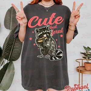 May include: Dark gray t-shirt with the text "Cute but feral" in pink. The shirt features a raccoon graphic eating a sandwich, surrounded by pink stars. The shirt is a casual style.