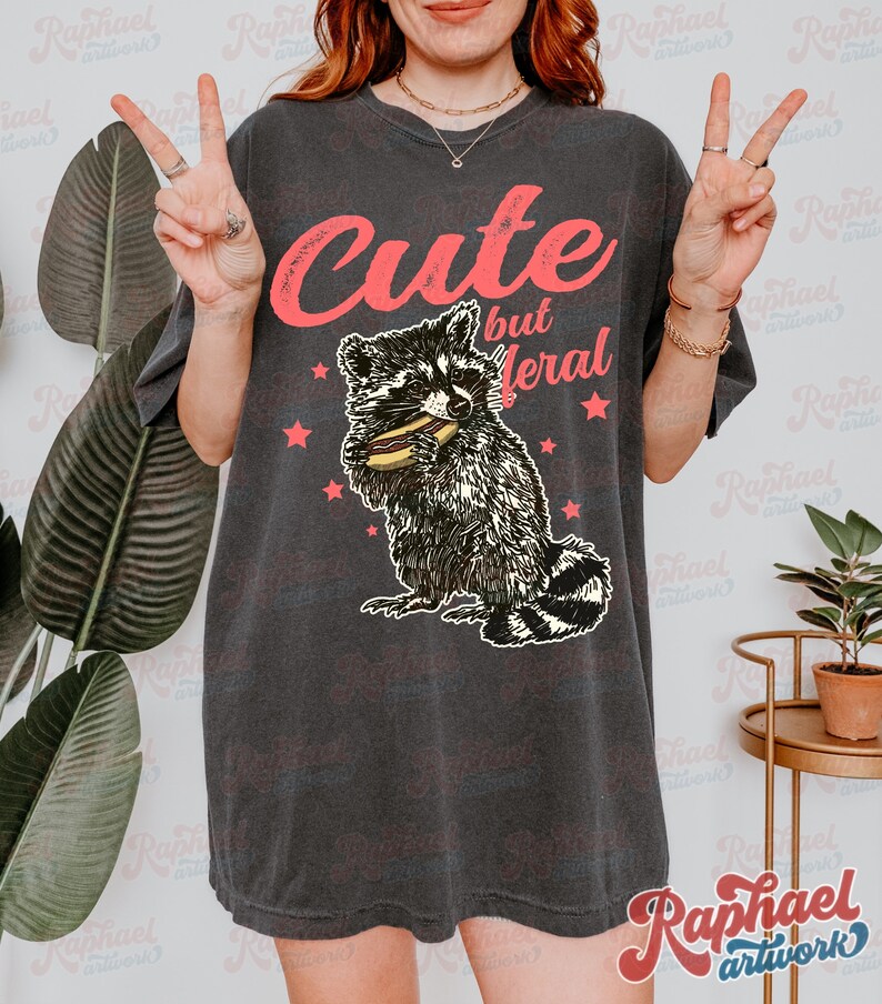 May include: Dark gray t-shirt with the text "Cute but feral" in pink. The shirt features a raccoon graphic eating a sandwich, surrounded by pink stars. The shirt is a casual style.