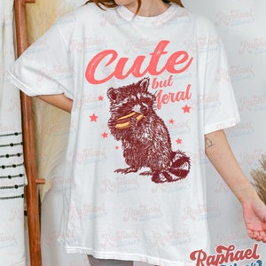 May include: White t-shirt featuring a raccoon illustration eating a hotdog, with the text "Cute but Feral" in red. The design includes red stars. The shirt is a casual, oversized style.