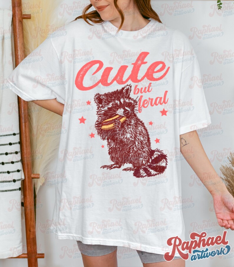 May include: White t-shirt featuring a raccoon illustration eating a hotdog, with the text "Cute but Feral" in red. The design includes red stars. The shirt is a casual, oversized style.