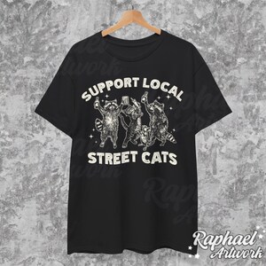May include: Black t-shirt with a white graphic of raccoons drinking beer and the text "Support Local Street Cats".