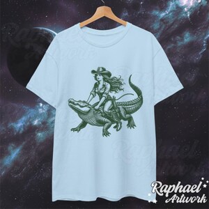 K&ouml;nnte beinhalten: Hellblaues T-Shirt mit einem grafischen Aufdruck einer Frau im Cowboyhut, die auf einem Alligator reitet.