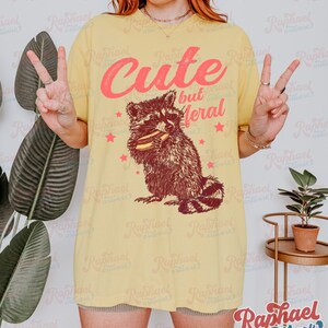 May include: Yellow t-shirt with a raccoon graphic holding a sandwich. The text "Cute but Meral" is above the raccoon in pink lettering. Red stars are scattered around the raccoon. The shirt is a casual style.