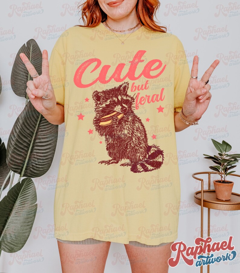 May include: Yellow t-shirt with a raccoon graphic holding a sandwich. The text "Cute but Meral" is above the raccoon in pink lettering. Red stars are scattered around the raccoon. The shirt is a casual style.