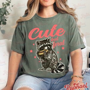 May include: Olive green t-shirt with the text "Cute but feral" in red. A raccoon graphic eating a hot dog is below the text, with red star accents. The shirt is a casual, unisex style.