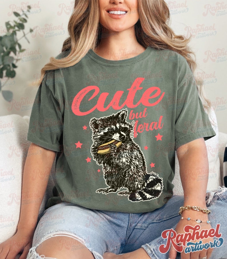 May include: Olive green t-shirt with the text "Cute but feral" in red. A raccoon graphic eating a hot dog is below the text, with red star accents. The shirt is a casual, unisex style.