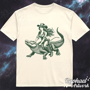 K&ouml;nnte beinhalten: Ein wei&szlig;es T-Shirt mit einer gr&uuml;nen Illustration einer Frau, die einen Cowboyhut tr&auml;gt und auf einem Alligator reitet.