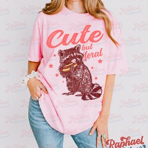 May include: Pink t-shirt with the text "Cute but Feral" above a raccoon graphic. The raccoon is brown and white, holding food. The shirt is worn with blue jeans.
