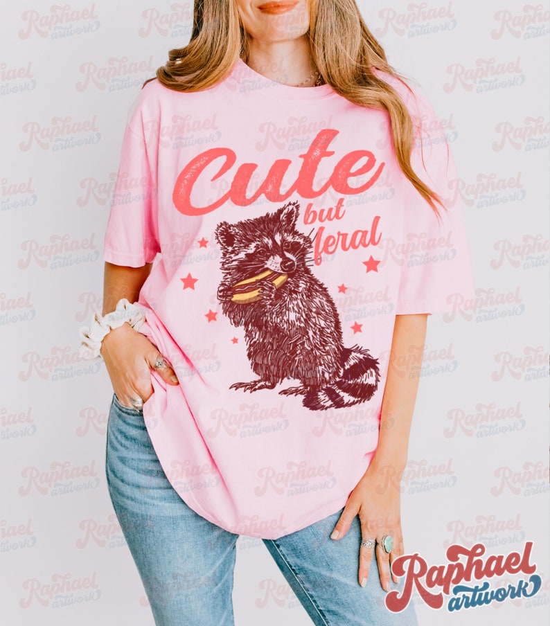 May include: Pink t-shirt with the text "Cute but Feral" above a raccoon graphic. The raccoon is brown and white, holding food. The shirt is worn with blue jeans.