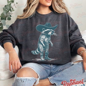 May include: A dark gray sweatshirt with a graphic of a raccoon wearing a cowboy hat and boots. The raccoon is standing on its hind legs and has a mischievous expression on its face.