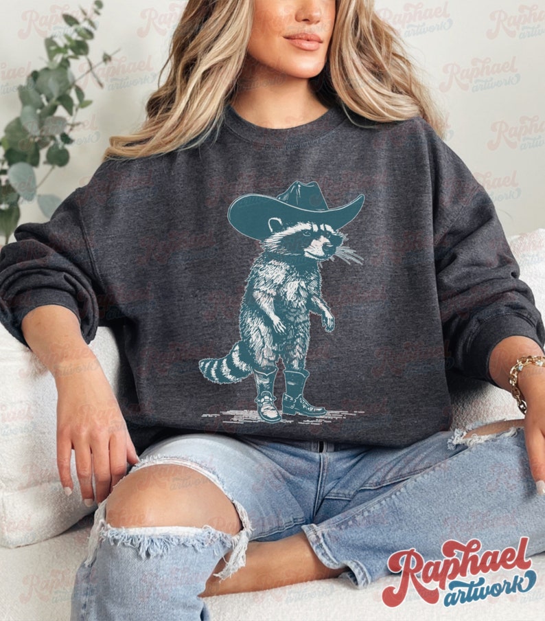May include: A dark gray sweatshirt with a graphic of a raccoon wearing a cowboy hat and boots. The raccoon is standing on its hind legs and has a mischievous expression on its face.