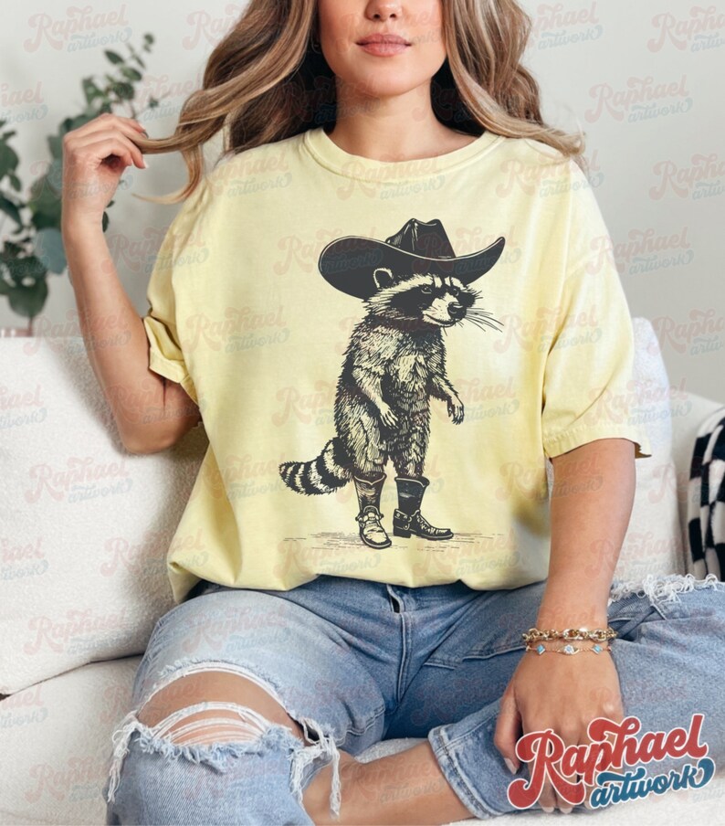 May include: A yellow t-shirt with a graphic of a raccoon wearing a cowboy hat and boots. The raccoon is standing on a light brown background.