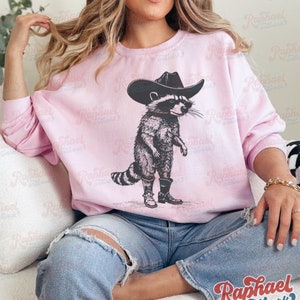 May include: A pink sweatshirt with a graphic of a raccoon wearing a cowboy hat and boots. The raccoon is standing on its hind legs and has a serious expression on its face.