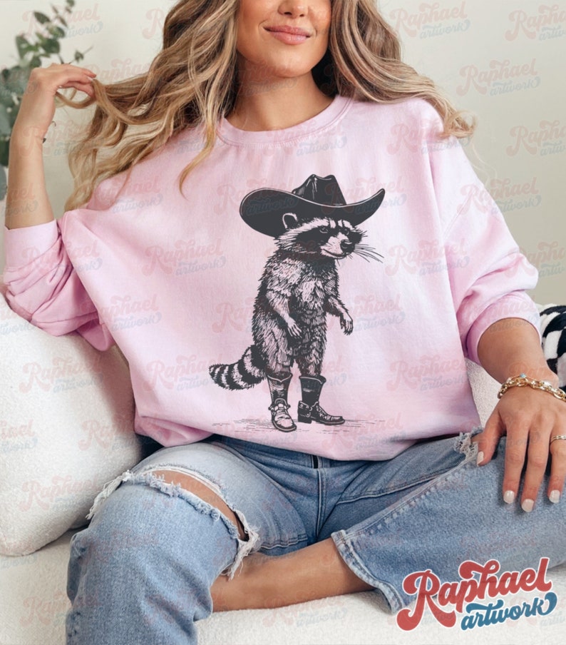 May include: A pink sweatshirt with a graphic of a raccoon wearing a cowboy hat and boots. The raccoon is standing on its hind legs and has a serious expression on its face.