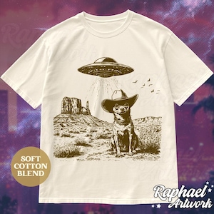 May include: A white t-shirt with a brown graphic of a chihuahua wearing a cowboy hat and standing in front of a desert landscape with a UFO hovering above. The text "Soft Cotton Blend" is in a brown rectangle in the lower left corner of the image. The text "Raphael Artwork" is in the lower right corner of the image.