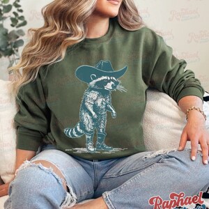 May include: A green sweatshirt with a graphic of a raccoon wearing a cowboy hat and boots. The raccoon is standing on its hind legs and has a mischievous expression on its face.