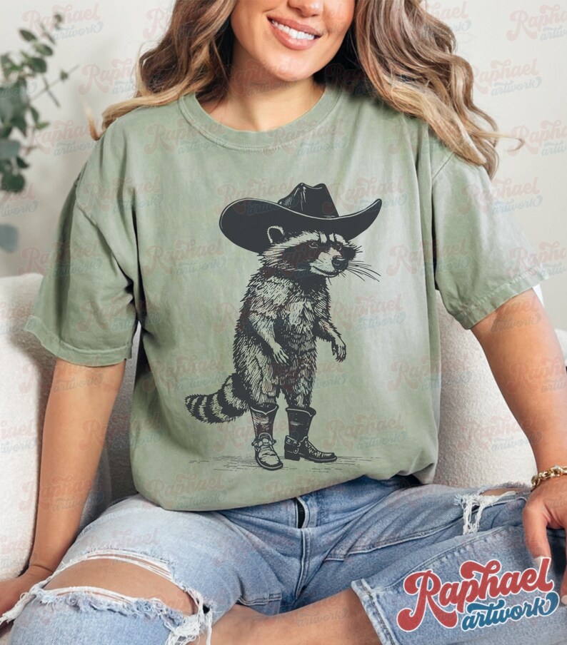 May include: A green t-shirt with a graphic of a raccoon wearing a cowboy hat and boots. The raccoon is standing on its hind legs and has a serious expression on its face.